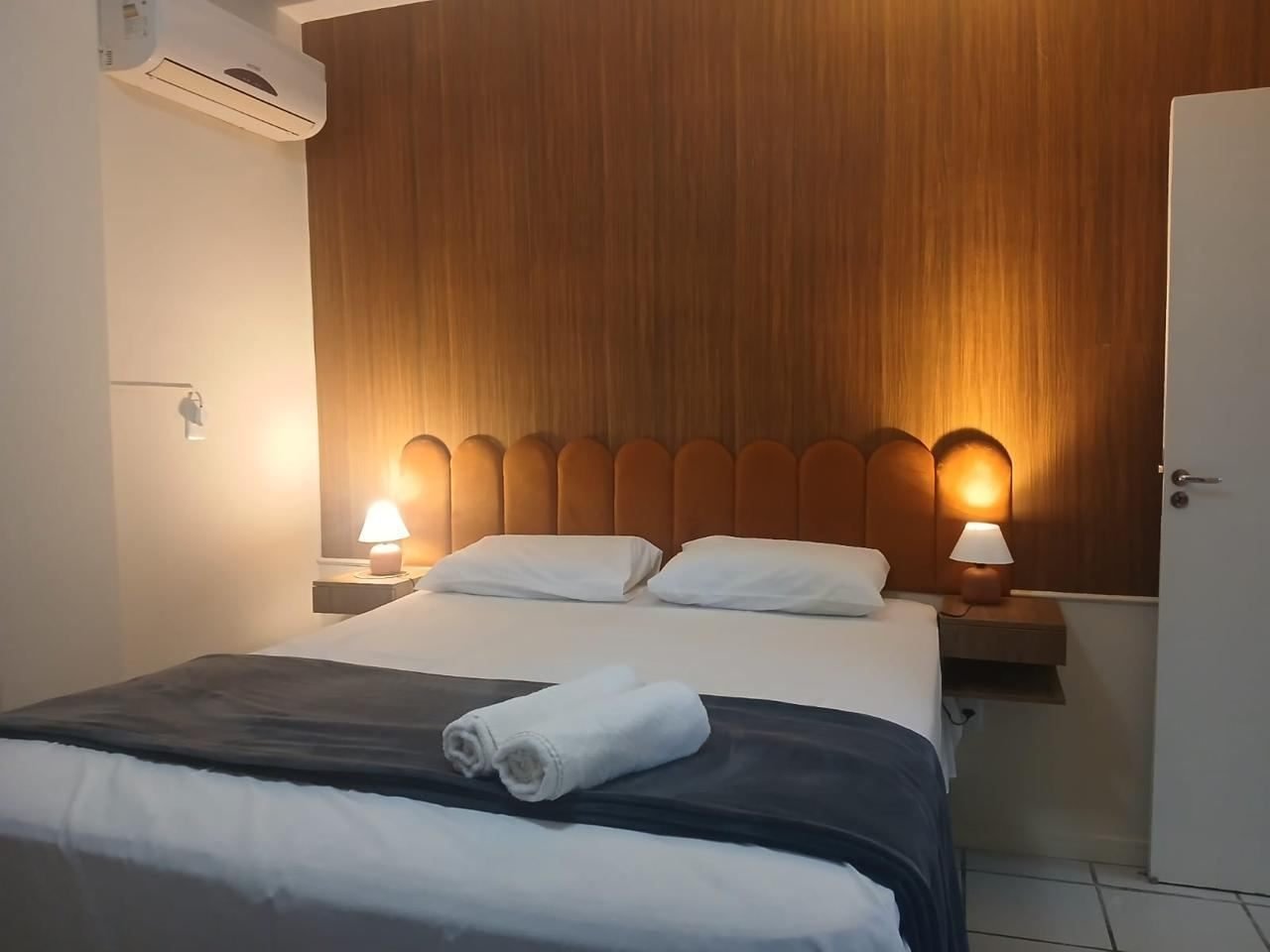 Quarto com cama de casal, cabeceira estofada em tom caramelo, abajures e ar-condicionado
