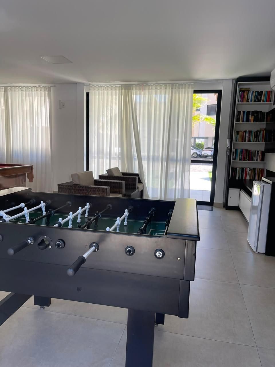 Sala de jogos do condomínio com mesa de pebolim, sinuca, poltronas e estante de livros
